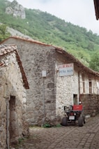 A rustic stone building with a red-tiled roof is situated in a mountainous, green area. A sign on the building reads 'Albergue Pe&ntilde;a Main'. An off-road vehicle or quad bike is parked nearby. The surroundings include cobblestone paths and lush greenery, evoking a tranquil, rural atmosphere.