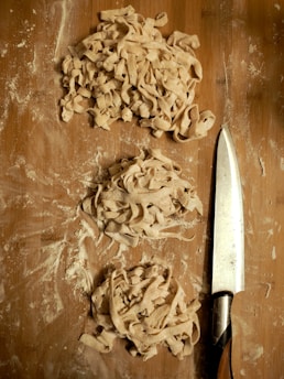 Piles of freshly cut pasta rest on a floured wooden surface, with a sharp knife placed nearby. The dough is rough and unevenly cut, suggesting a homemade preparation process.