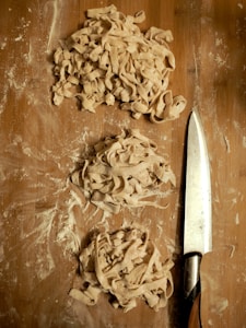 Piles of freshly cut pasta rest on a floured wooden surface, with a sharp knife placed nearby. The dough is rough and unevenly cut, suggesting a homemade preparation process.