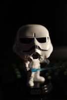 A small, stylized figurine of a character in white armor with a black visor and features resembling a helmet. The figure is holding a blue lightsaber and stands against a dark background with subtle green bokeh effects.