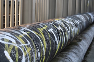 A large industrial pipe runs horizontally alongside a series of vertical metal bars. The pipe is covered in graffiti with prominent white and yellow markings. The surrounding environment appears to be urban or industrial.