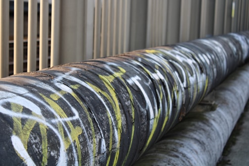 A large industrial pipe runs horizontally alongside a series of vertical metal bars. The pipe is covered in graffiti with prominent white and yellow markings. The surrounding environment appears to be urban or industrial.
