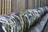 A large industrial pipe runs horizontally alongside a series of vertical metal bars. The pipe is covered in graffiti with prominent white and yellow markings. The surrounding environment appears to be urban or industrial.