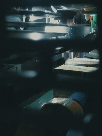 A dimly lit industrial or mechanical setting showing parts of machinery or equipment with a focus on metal and shadowed components. Some light reflects off surfaces, creating a contrasting interplay between dark and light sections.