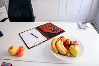 A clean, organized desk featuring a white surface. On the desk, there is a white bowl filled with assorted fruits such as bananas, apples, and an orange. Nearby, a clipboard with a calendar or schedule is placed next to a pen. There is also a brown folder or case containing a calculator, and a separate calculator can be seen further on the desk. An office chair is positioned behind the desk.