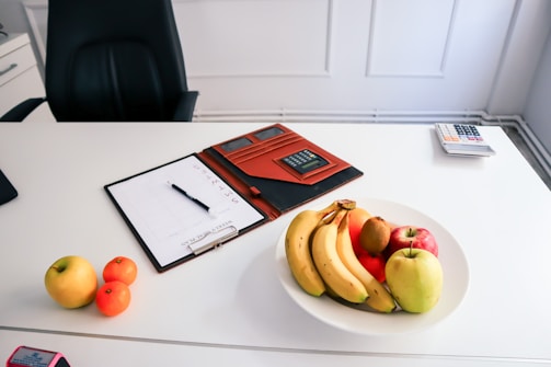 A clean, organized desk featuring a white surface. On the desk, there is a white bowl filled with assorted fruits such as bananas, apples, and an orange. Nearby, a clipboard with a calendar or schedule is placed next to a pen. There is also a brown folder or case containing a calculator, and a separate calculator can be seen further on the desk. An office chair is positioned behind the desk.