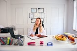 A person with long hair is sitting at a desk in a brightly lit office environment. They are writing on a clipboard and surrounded by various office supplies including a laptop, pens, and a stapler. On the desk, there is also a bowl of fruit and some stacked books. Certificates or diplomas are framed on the wall behind.