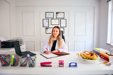 A person with long hair is sitting at a desk in a brightly lit office environment. They are writing on a clipboard and surrounded by various office supplies including a laptop, pens, and a stapler. On the desk, there is also a bowl of fruit and some stacked books. Certificates or diplomas are framed on the wall behind.
