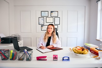 A person with long hair is sitting at a desk in a brightly lit office environment. They are writing on a clipboard and surrounded by various office supplies including a laptop, pens, and a stapler. On the desk, there is also a bowl of fruit and some stacked books. Certificates or diplomas are framed on the wall behind.