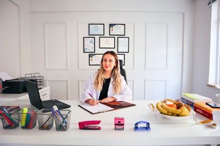 A person with long hair is sitting at a desk in a brightly lit office environment. They are writing on a clipboard and surrounded by various office supplies including a laptop, pens, and a stapler. On the desk, there is also a bowl of fruit and some stacked books. Certificates or diplomas are framed on the wall behind.