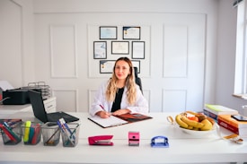 A person with long hair is sitting at a desk in a brightly lit office environment. They are writing on a clipboard and surrounded by various office supplies including a laptop, pens, and a stapler. On the desk, there is also a bowl of fruit and some stacked books. Certificates or diplomas are framed on the wall behind.