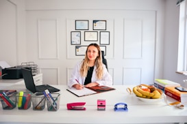 A person with long hair is sitting at a desk in a brightly lit office environment. They are writing on a clipboard and surrounded by various office supplies including a laptop, pens, and a stapler. On the desk, there is also a bowl of fruit and some stacked books. Certificates or diplomas are framed on the wall behind.