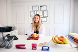A woman in a white coat is sitting at a desk, holding an orange. Behind her, there are framed certificates on the wall. The desk has various items including a laptop, a basket of office supplies, a stapler, a hole punch, a plate of fruit including bananas and apples, and some books. A measuring tape is draped across the desk.