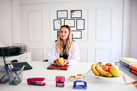 A woman in a white coat is sitting at a desk, holding an orange. Behind her, there are framed certificates on the wall. The desk has various items including a laptop, a basket of office supplies, a stapler, a hole punch, a plate of fruit including bananas and apples, and some books. A measuring tape is draped across the desk.