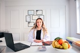 A person in a professional setting is seated at a desk with a white coat on. There is a laptop to the left and a clipboard with papers in front. A bowl of fresh fruits including bananas and apples is on the right side of the desk. Several certificates or diplomas are hung on the wall in the background.