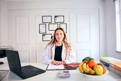 A person in a professional setting is seated at a desk with a white coat on. There is a laptop to the left and a clipboard with papers in front. A bowl of fresh fruits including bananas and apples is on the right side of the desk. Several certificates or diplomas are hung on the wall in the background.