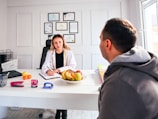 A professional consultation setting with a medical professional sitting at a desk facing a client. The room has a modern aesthetic with white walls decorated with framed certificates. The desk is organized with office supplies, a laptop, and a fruit bowl in the center.