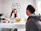A professional consultation setting with a medical professional sitting at a desk facing a client. The room has a modern aesthetic with white walls decorated with framed certificates. The desk is organized with office supplies, a laptop, and a fruit bowl in the center.