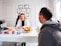 A professional consultation setting with a medical professional sitting at a desk facing a client. The room has a modern aesthetic with white walls decorated with framed certificates. The desk is organized with office supplies, a laptop, and a fruit bowl in the center.