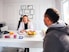 A professional consultation setting with a medical professional sitting at a desk facing a client. The room has a modern aesthetic with white walls decorated with framed certificates. The desk is organized with office supplies, a laptop, and a fruit bowl in the center.