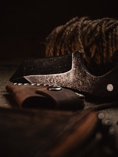 A close-up of a rugged, metallic blade with a weathered appearance, accompanied by a leather sheath. In the background, thick coiled rope adds to the rustic aesthetic.