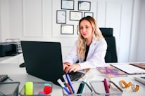 A woman in a white coat is seated at a desk working on a laptop. The desk is organized with various office supplies such as pens, notebooks, and a measuring tape. Certificates are neatly hung on the wall behind her, indicating a professional environment.