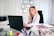 A woman in a white coat is seated at a desk working on a laptop. The desk is organized with various office supplies such as pens, notebooks, and a measuring tape. Certificates are neatly hung on the wall behind her, indicating a professional environment.