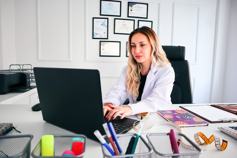 A woman in a white coat is seated at a desk working on a laptop. The desk is organized with various office supplies such as pens, notebooks, and a measuring tape. Certificates are neatly hung on the wall behind her, indicating a professional environment.