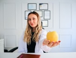 A woman with long, wavy blonde hair and a white lab coat is sitting at a desk. She holds a yellow apple in her outstretched hand. Behind her, a wall displays multiple certificates or diplomas in black frames. The setting appears to be a professional office or consultation room.