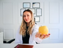 A woman with long, wavy blonde hair and a white lab coat is sitting at a desk. She holds a yellow apple in her outstretched hand. Behind her, a wall displays multiple certificates or diplomas in black frames. The setting appears to be a professional office or consultation room.