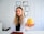A woman with long, wavy blonde hair and a white lab coat is sitting at a desk. She holds a yellow apple in her outstretched hand. Behind her, a wall displays multiple certificates or diplomas in black frames. The setting appears to be a professional office or consultation room.