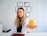 A woman with long, wavy blonde hair and a white lab coat is sitting at a desk. She holds a yellow apple in her outstretched hand. Behind her, a wall displays multiple certificates or diplomas in black frames. The setting appears to be a professional office or consultation room.