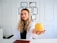 A woman with long, wavy blonde hair and a white lab coat is sitting at a desk. She holds a yellow apple in her outstretched hand. Behind her, a wall displays multiple certificates or diplomas in black frames. The setting appears to be a professional office or consultation room.