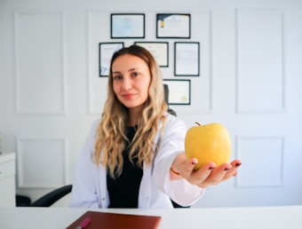 A woman with long, wavy blonde hair and a white lab coat is sitting at a desk. She holds a yellow apple in her outstretched hand. Behind her, a wall displays multiple certificates or diplomas in black frames. The setting appears to be a professional office or consultation room.