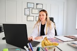 A person in a white lab coat is seated at a desk with a laptop. Several certificates or degrees are mounted on the wall behind them. A bunch of bananas and office supplies can be seen on the desk, along with notebooks or files.
