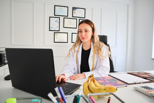 A person in a white lab coat is seated at a desk with a laptop. Several certificates or degrees are mounted on the wall behind them. A bunch of bananas and office supplies can be seen on the desk, along with notebooks or files.