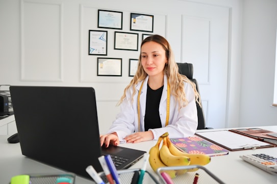 A person in a white lab coat is seated at a desk with a laptop. Several certificates or degrees are mounted on the wall behind them. A bunch of bananas and office supplies can be seen on the desk, along with notebooks or files.