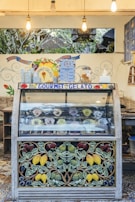 A gelato display case with a colorful and artistic design featuring fruit motifs. The display includes various flavors of gelato in round containers, each with a label. Above the case, there are hanging light bulbs and decorative signage with the words 'Gourmet Gelato'. A menu board with prices is visible, along with additional decoration on the walls.