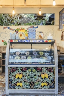 A gelato display case with a colorful and artistic design featuring fruit motifs. The display includes various flavors of gelato in round containers, each with a label. Above the case, there are hanging light bulbs and decorative signage with the words 'Gourmet Gelato'. A menu board with prices is visible, along with additional decoration on the walls.