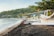Traditional fishing boats with colorful sails are lined up along a sandy beach. The shoreline curves towards lush, green hills in the background. The sea is calm, with gentle waves lapping at the dark sand. Trees and foliage line the edge of the beach, adding to the natural scenery.