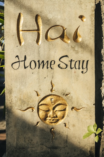 A textured stone wall features an embossed golden sun with a serene face and elaborate rays. Above the sun, the words 'Hai Home Stay' are artistically written, with 'Hai' in large, cursive golden letters and 'Home Stay' in smaller black script. A small green plant leaf appears in the bottom right corner.