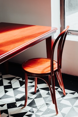 A wooden chair with a curved back rests against a wooden table, both illuminated by sunlight streaming in from a nearby window. The floor features a geometric black and white tile pattern, adding contrast to the warm wood tones of the furniture.