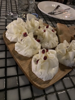 A wooden board features several dumpling-like items topped with a few red pomegranate seeds, placed on a black and white tiled table. Next to them, a piece of bread rests on the board. In the background, there are table settings including a glass, a plate, and cutlery.