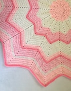 A crocheted blanket featuring a zigzag pattern with alternating shades of pink and cream. The design includes starburst motifs and textured stitching, creating a visually appealing contrast.