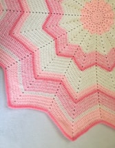 A crocheted blanket featuring a zigzag pattern with alternating shades of pink and cream. The design includes starburst motifs and textured stitching, creating a visually appealing contrast.