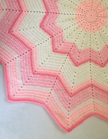 A crocheted blanket featuring a zigzag pattern with alternating shades of pink and cream. The design includes starburst motifs and textured stitching, creating a visually appealing contrast.