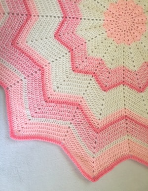 A crocheted blanket featuring a zigzag pattern with alternating shades of pink and cream. The design includes starburst motifs and textured stitching, creating a visually appealing contrast.