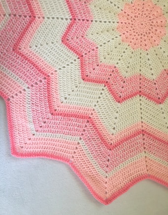 A crocheted blanket featuring a zigzag pattern with alternating shades of pink and cream. The design includes starburst motifs and textured stitching, creating a visually appealing contrast.