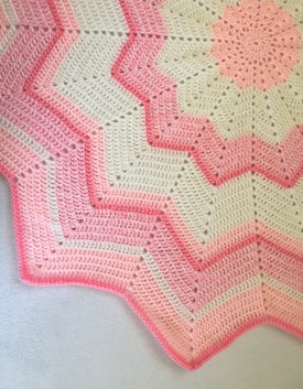 A crocheted blanket featuring a zigzag pattern with alternating shades of pink and cream. The design includes starburst motifs and textured stitching, creating a visually appealing contrast.