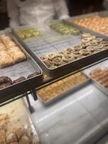 Several trays of various types of desserts are on display, including pastries with visible nuts and powdered sugar. The background shows more trays of similar desserts, creating a vibrant and appetizing arrangement.